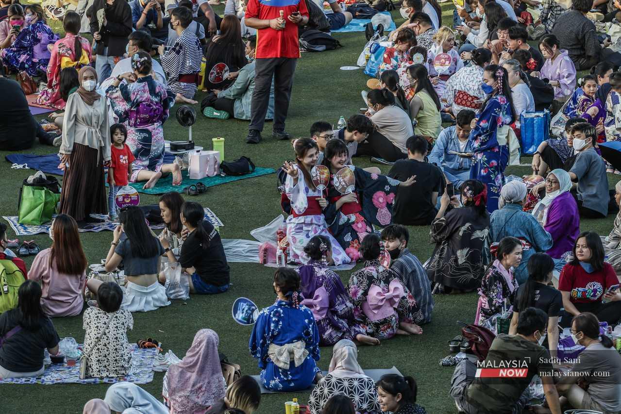 Laughing and chatting, participants, many dressed in traditional Japanese outfits, take pictures on blankets spread out on the grass before the start of the festival. 
