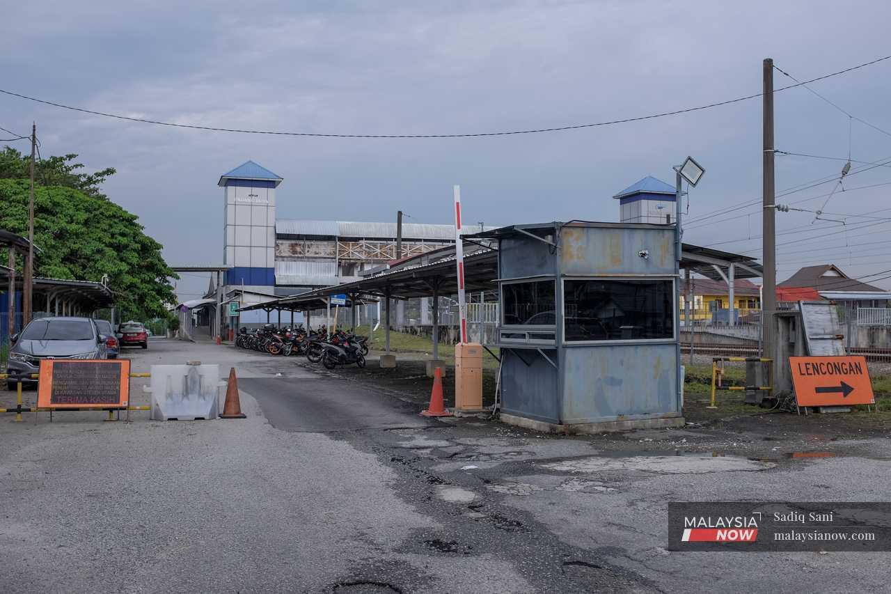 The road at the entrance to the Padang Jawa station is riddled with potholes, with cars parked along the side and traffic cones used as part of the barrier. 