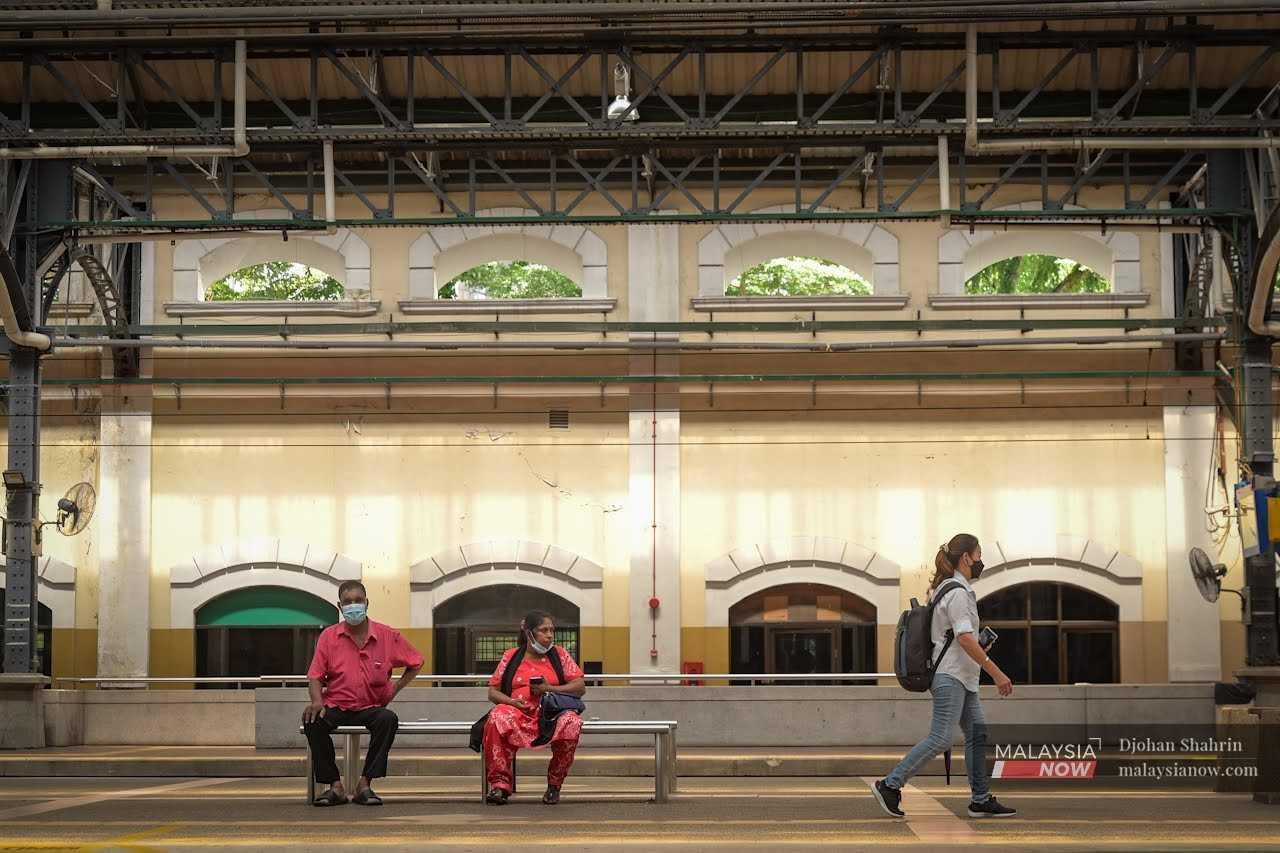 Dua penumpang menunggu di atas bangku sementara seorang lagi wanita sedang berjalan mencari tempat duduk di platform.