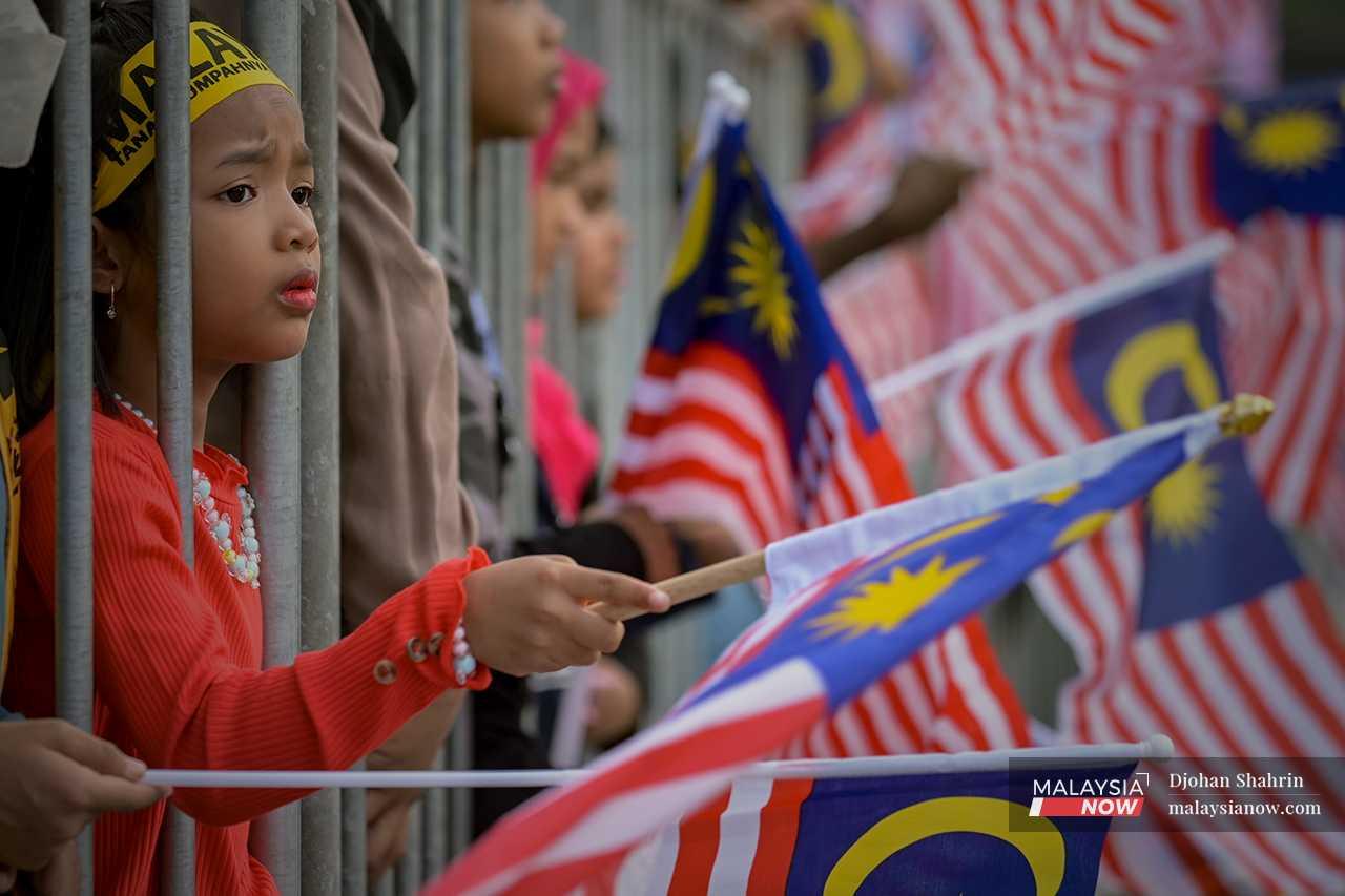A child waves the Jalur Gemilang as she watches the parade through the metal divider. 