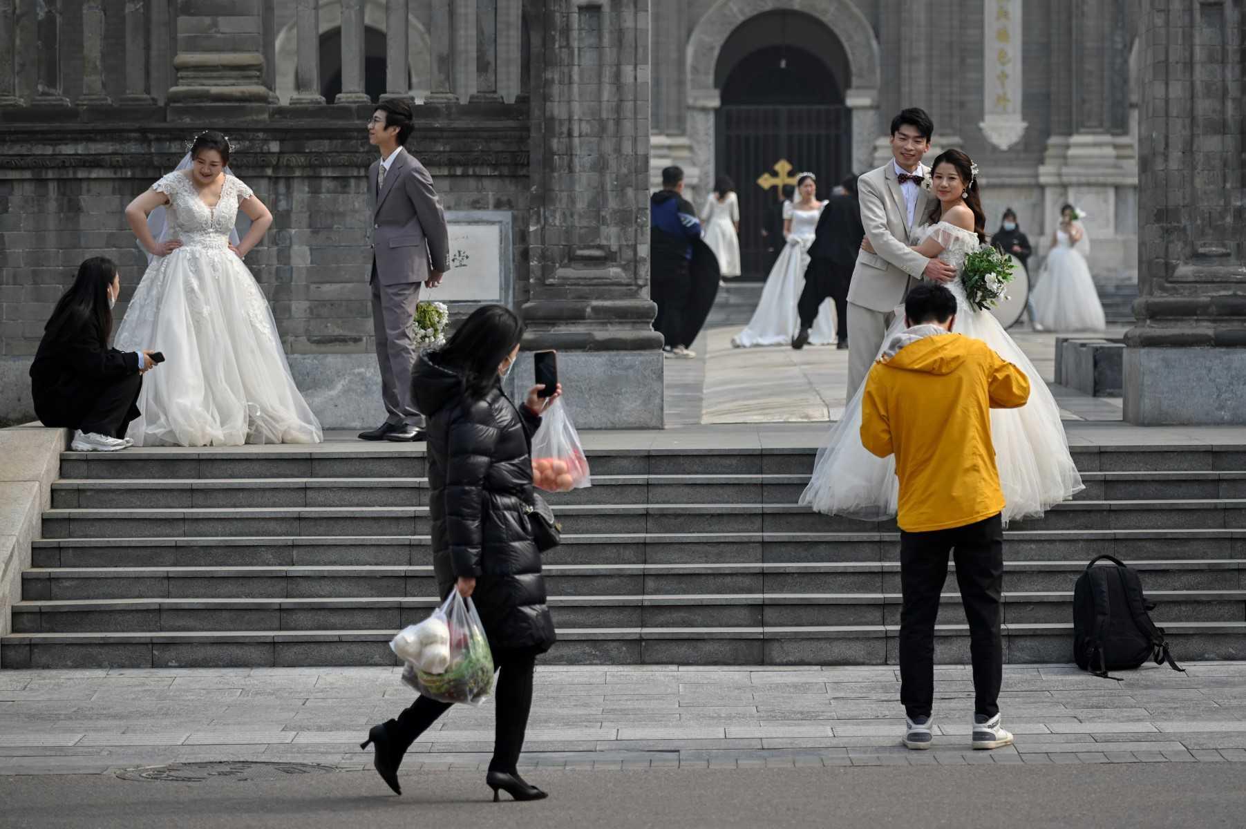 A woman uses her mobile phone to take a picture while walking past a group of couples taking wedding pictures in front of a church in Beijing on March 11, 2021. Photo: AFP