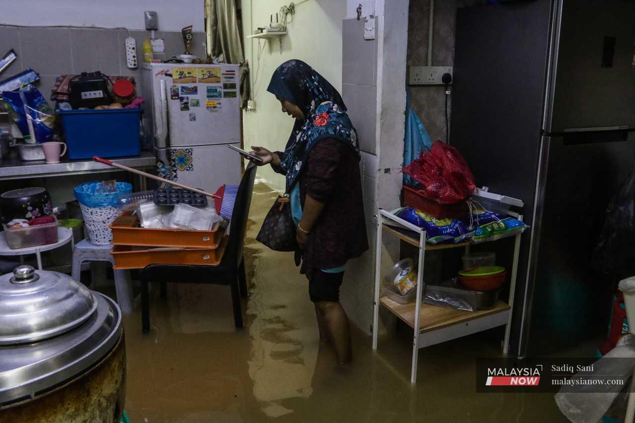 A woman checks her phone as she stands with her pants rolled up in her waterlogged kitchen. She says she may have lost as much as RM2,000 in appliances due to the flood. 