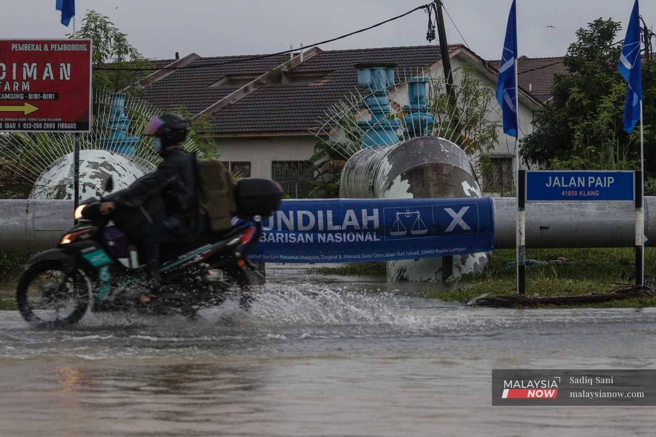 Seorang penunggang motorsikal melewati deretan bendera parti di jalan yang dinaiki air banjir.