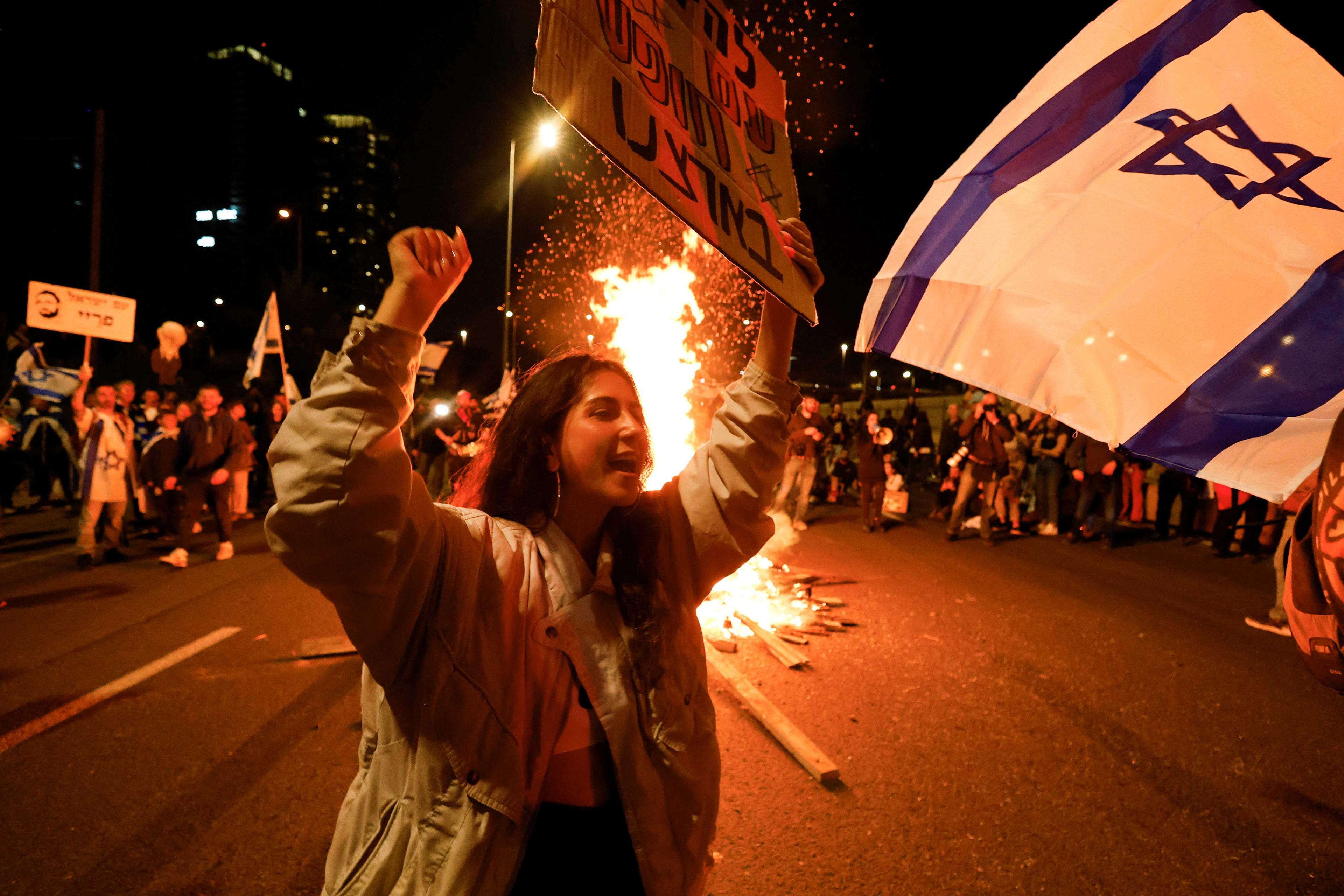 Seorang wanita mengangkat sepanduk di sebelah unggun api di tengah jalan raya ketika protes terhadap kerajaan pimpinan Benjamin Netanyahu dan cadangan pembaharuan kehakimannya untuk mengurangkan kuasa Mahkamah Agung di Tel Aviv, Israel, 25 Februari. Gambar: Reuters