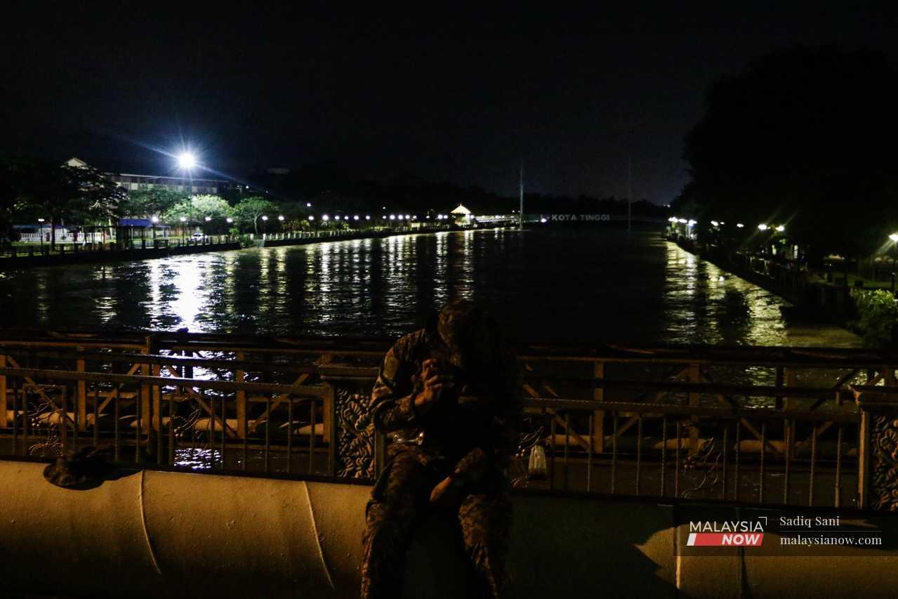 An army personnel talks on the phone as the banks of Sungai Johor loom behind. 