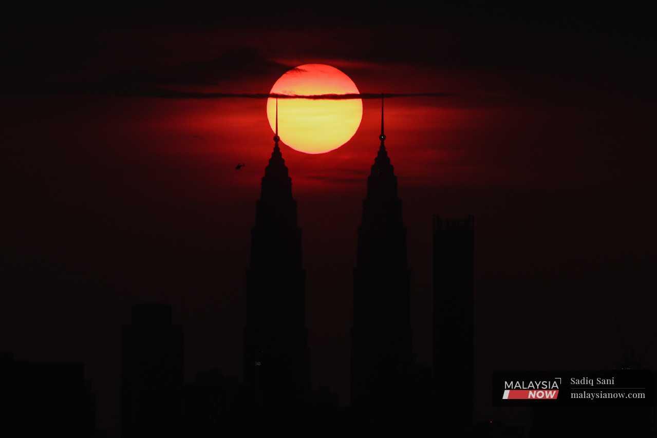 Sebuah helikopter melintasi Menara Berkembar Petronas, berlatarbelakangkan matahari terbenam.