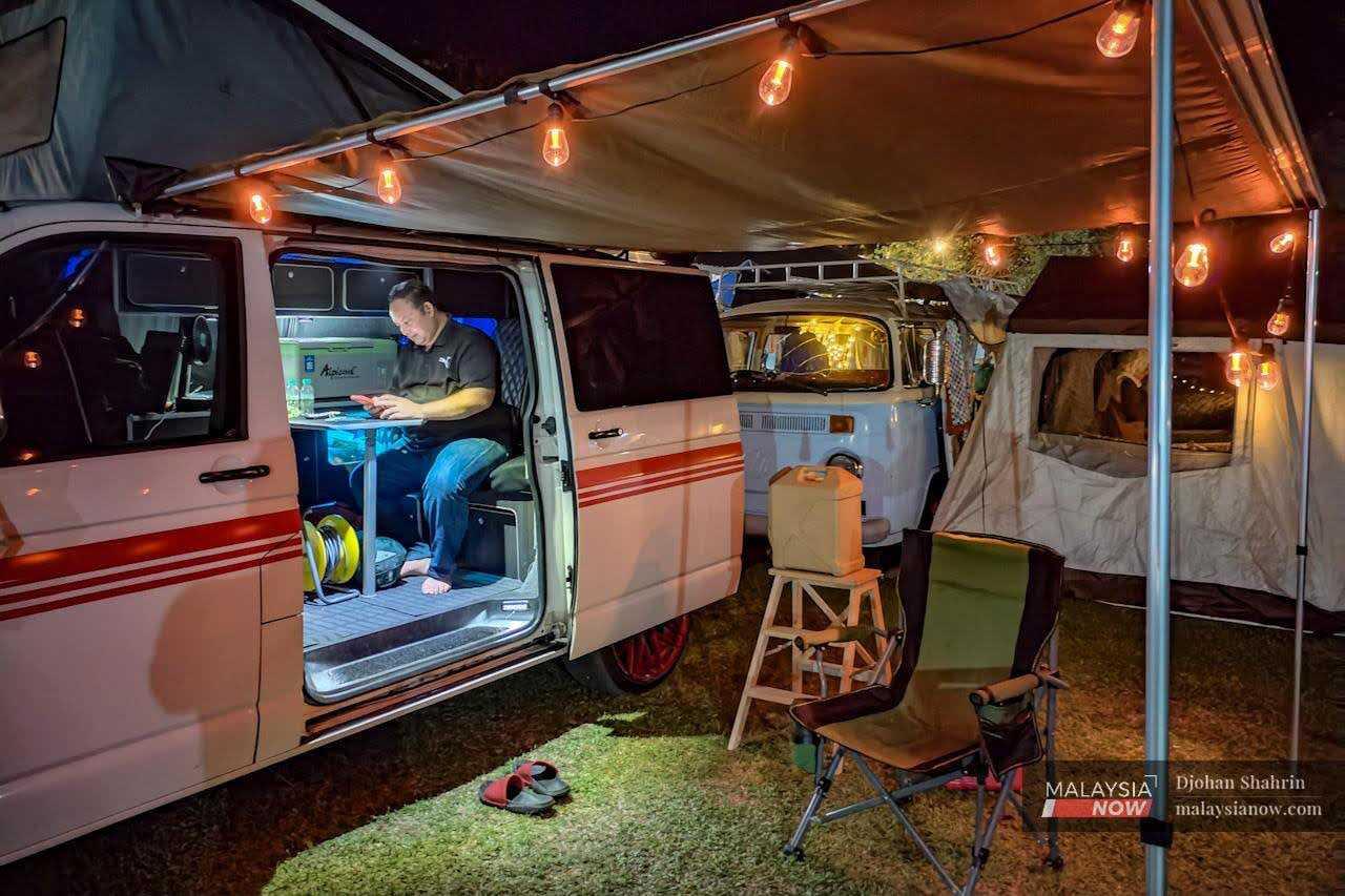 A man uses his phone in his Kombi caravan, which is hung about with lights. 