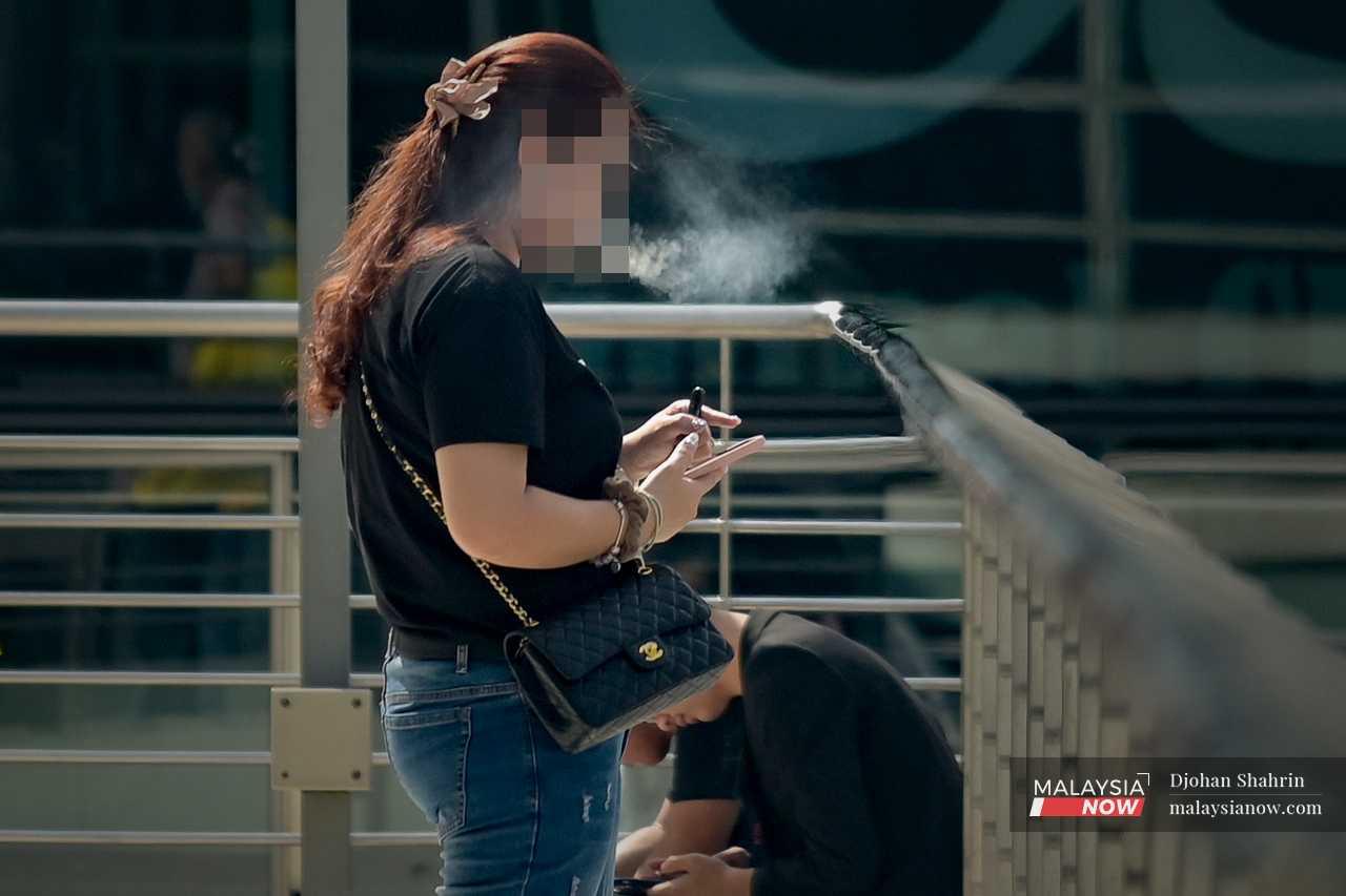 Seorang wanita menghisap vape sambil menggunakan telefonnya di lapangan terbang. 