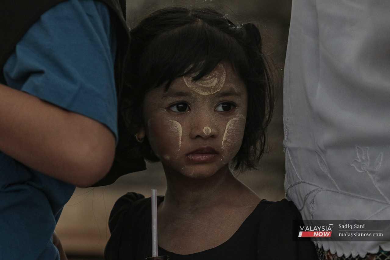 A young girl with powder on her face watches from behind her mother.