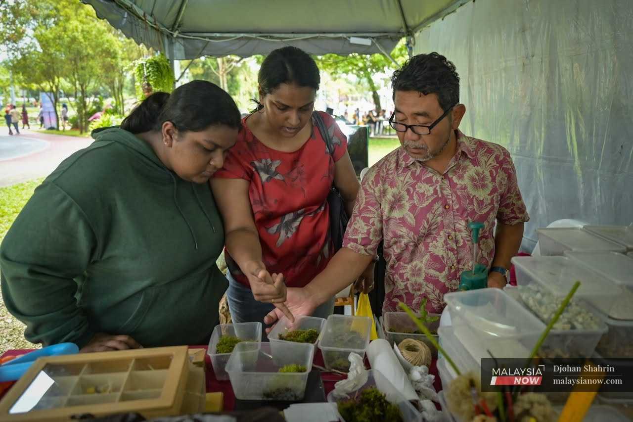 Two customers examine the moss being sold in containers as Aziz provides tips on how to care for the Octoblepharum moss.