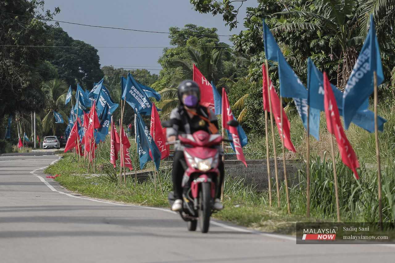 Seorang penunggang motosikal melintasi deretan bendera Perikatan Nasional dan Pakatan Harapan yang dipacak di tepi jalan menjelang pilihan raya kecil Simpang Jeram.