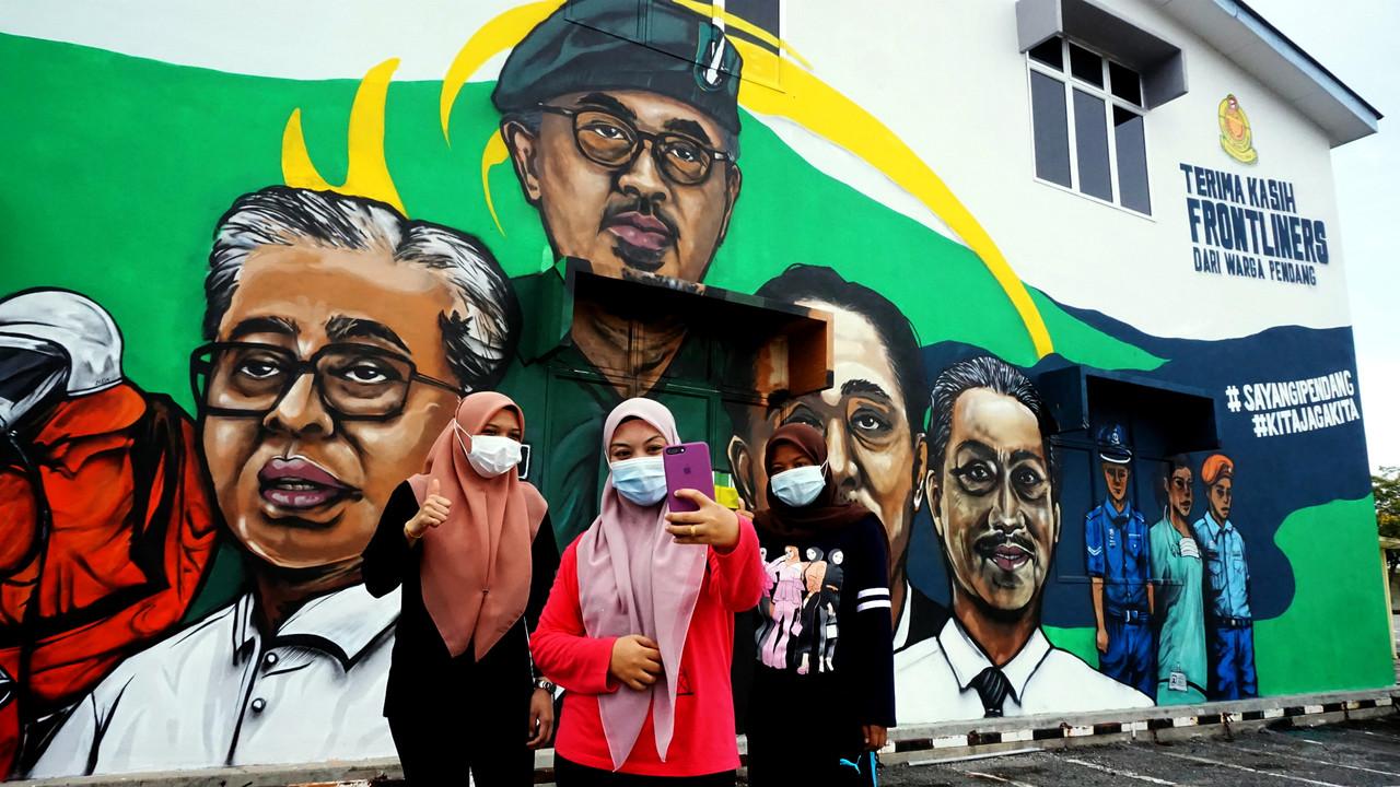 Penduduk Pendang, Anis Atikah Ismail, 22, (tengah) ditemani rakannya, Ayu Wahida Abdul Hadis, 27, (kanan) dan Farhana Mohd Nor, 24, (kiri) berswafoto di hadapan mural Majlis Daerah Pendang (MDP) bagi menghargai barisan frontliner yang bertungkus lumus memerangi pandemik Covid-19 di negara ini di pekan Pendang.Antara potret dipaparkan ialah Yang di-Pertuan Agong Al-Sultan Abdullah Ri'ayatuddin Al-Mustafa Billah Shah, Perdana Menteri Muhyiddin Yassin, Menteri Kanan (Kluster Keselamatan) Ismail Sabri Yaakob, Ketua Pengarah Kesihatan Dr Noor Hisham Abdullah serta barisan anggota keselamatan yang terlibat. Gambar: Bernama