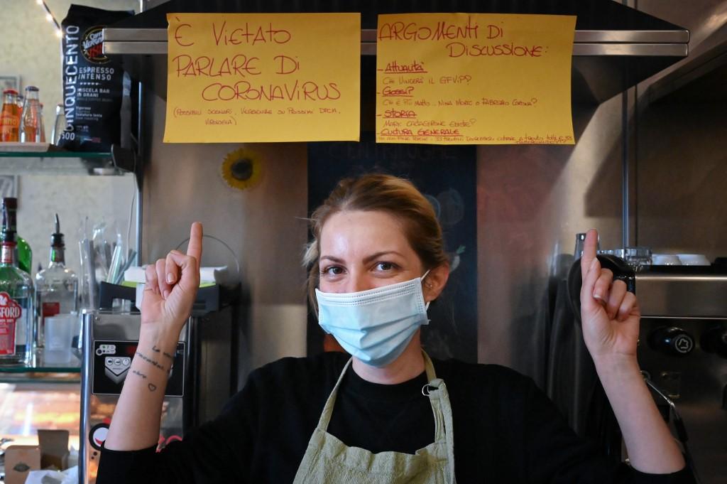 Bar owner Cristina Mattioli poses by a sign (top left) reading: 'It is forbidden to talk about the coronavirus' and another suggesting other topics of conversation instead. Photo: AFP