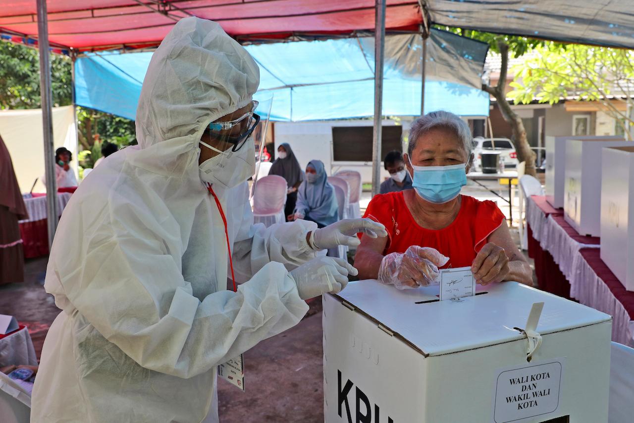 An electoral worker wearing a protective suit assists an elderly woman in casting her ballot during the regional election at a polling station in Tangerang, Indonesia, Dec 9. Photo: AP