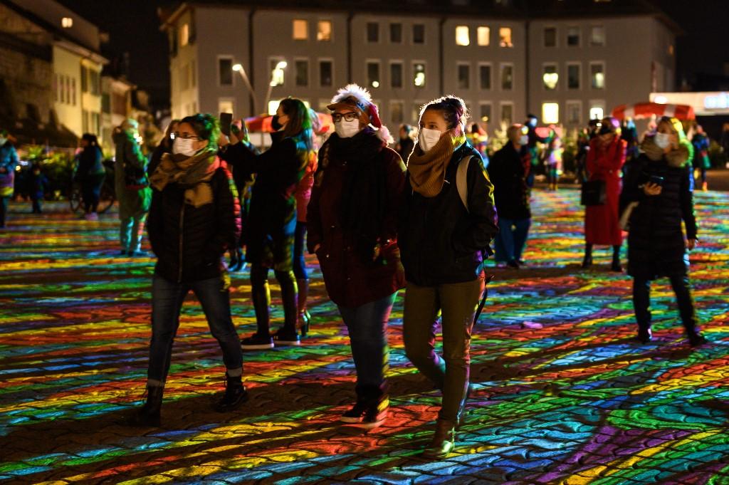 People wearing face masks walk at the Place de la Riponne square illuminated during a lightshow in downtown Lausanne, Dec 17. Photo: AFP