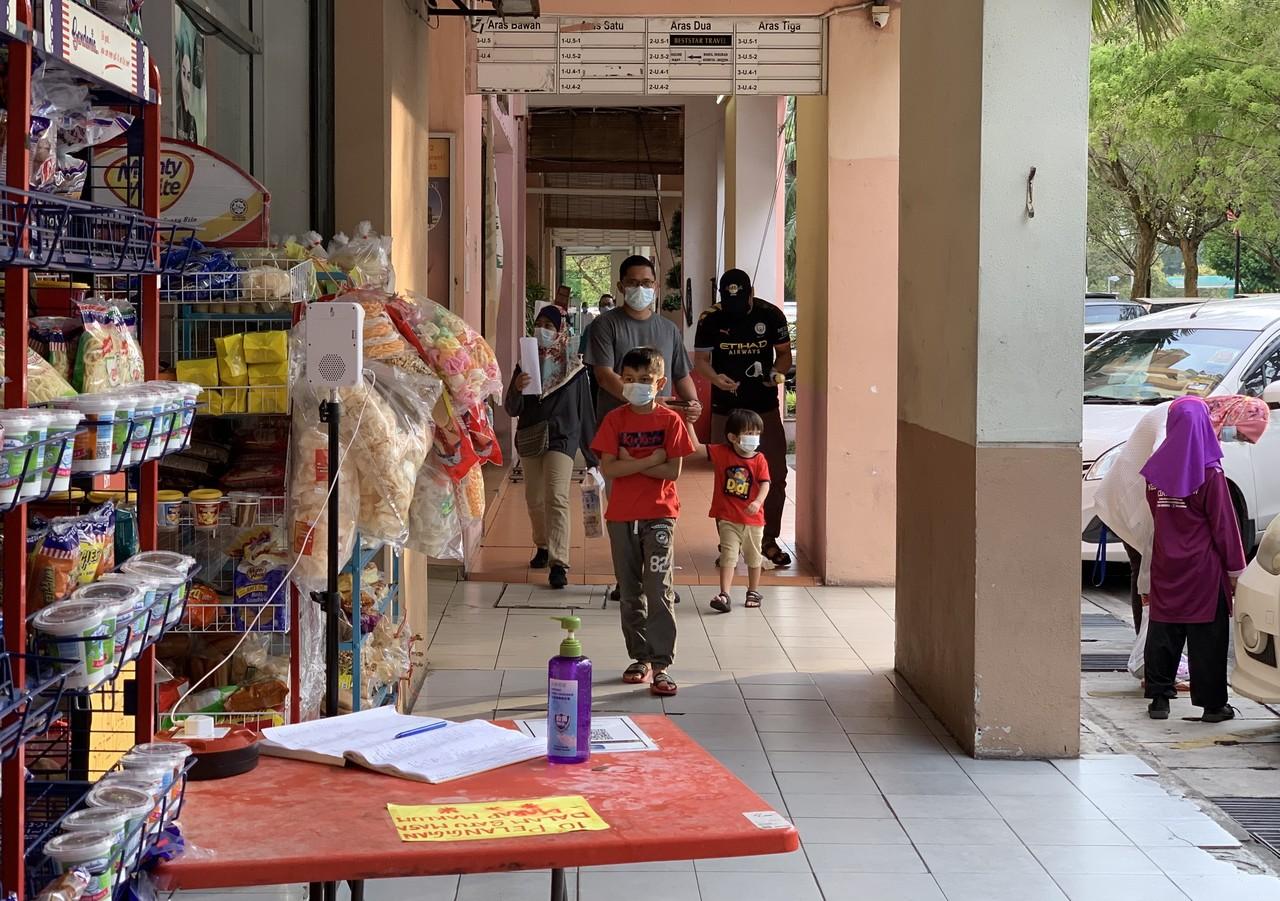 People walk past a sundry shop in Putrajaya where hand sanitiser, a log book and the MySejahtera QR code are laid out on a table alongside a thermometer for temperature checks. Photo: Bernama