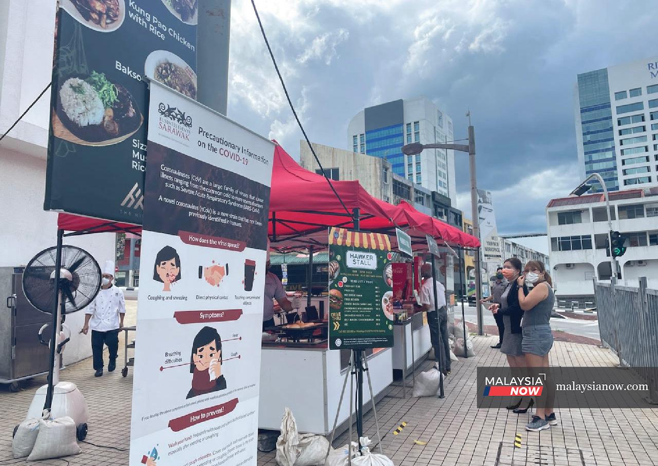 Lunch-hour customers wait for their food to be packed at Pullman Kuching's hawker stall located directly outside the hotel building. Posters and signs remind them to maintain a safe distance and to check in using the MySejahtera app.