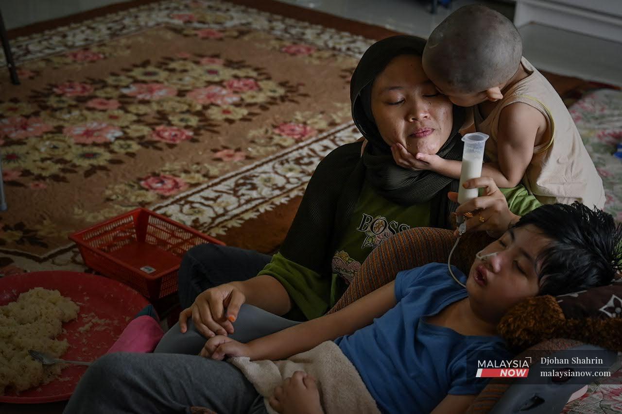 Zaitul Akma Muhd Zin feeds her daughter, Aliya Darwisyah, milk through a tube in her nose while her son, Adib Aisy Rizqi, leans over to give her a kiss.