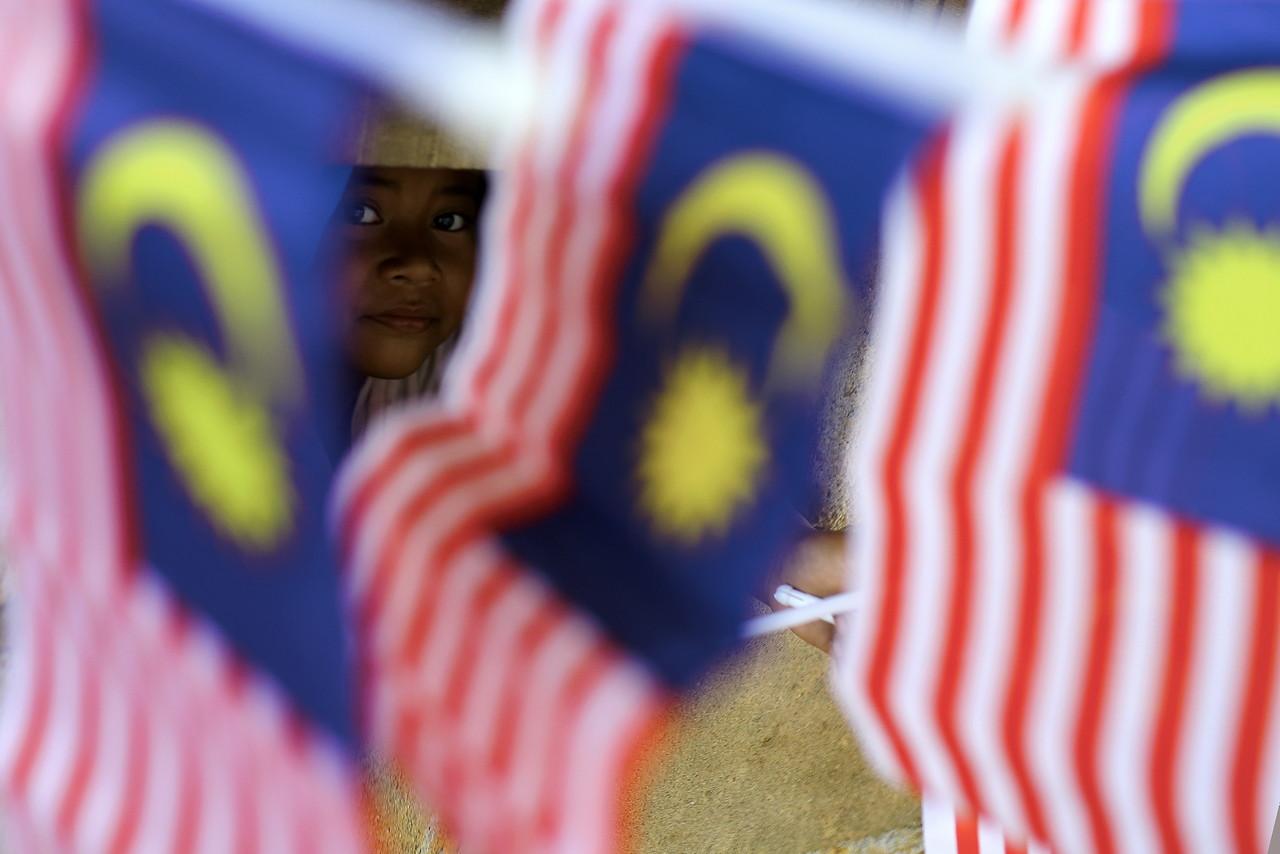 An Orang Asli child from the Semelai community looks out from behind Malaysian flags, put up at her home ahead of Merdeka Day celebrations this month. Photo: Bernama
