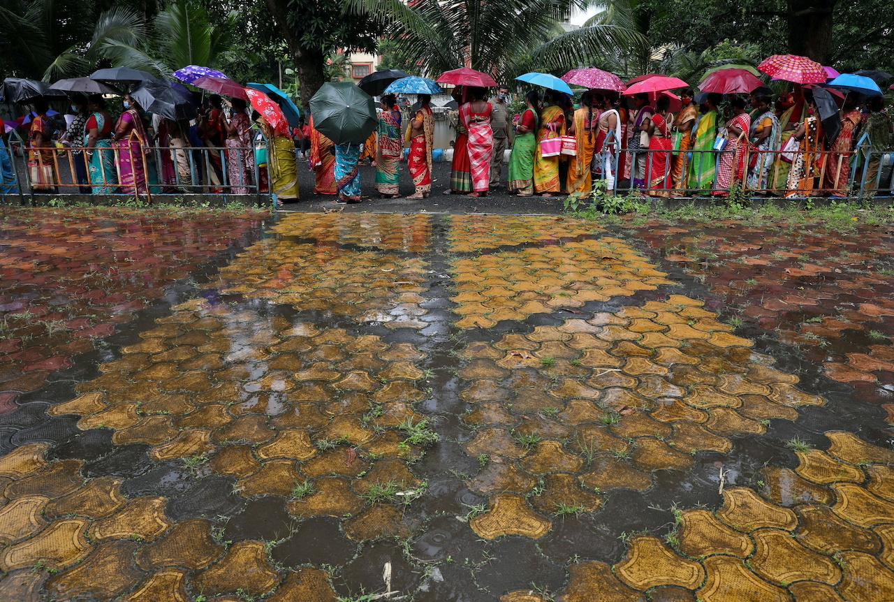 Women hold umbrellas to cover from rain as they wait to receive a dose of COVISHIELD vaccine in Kolkata