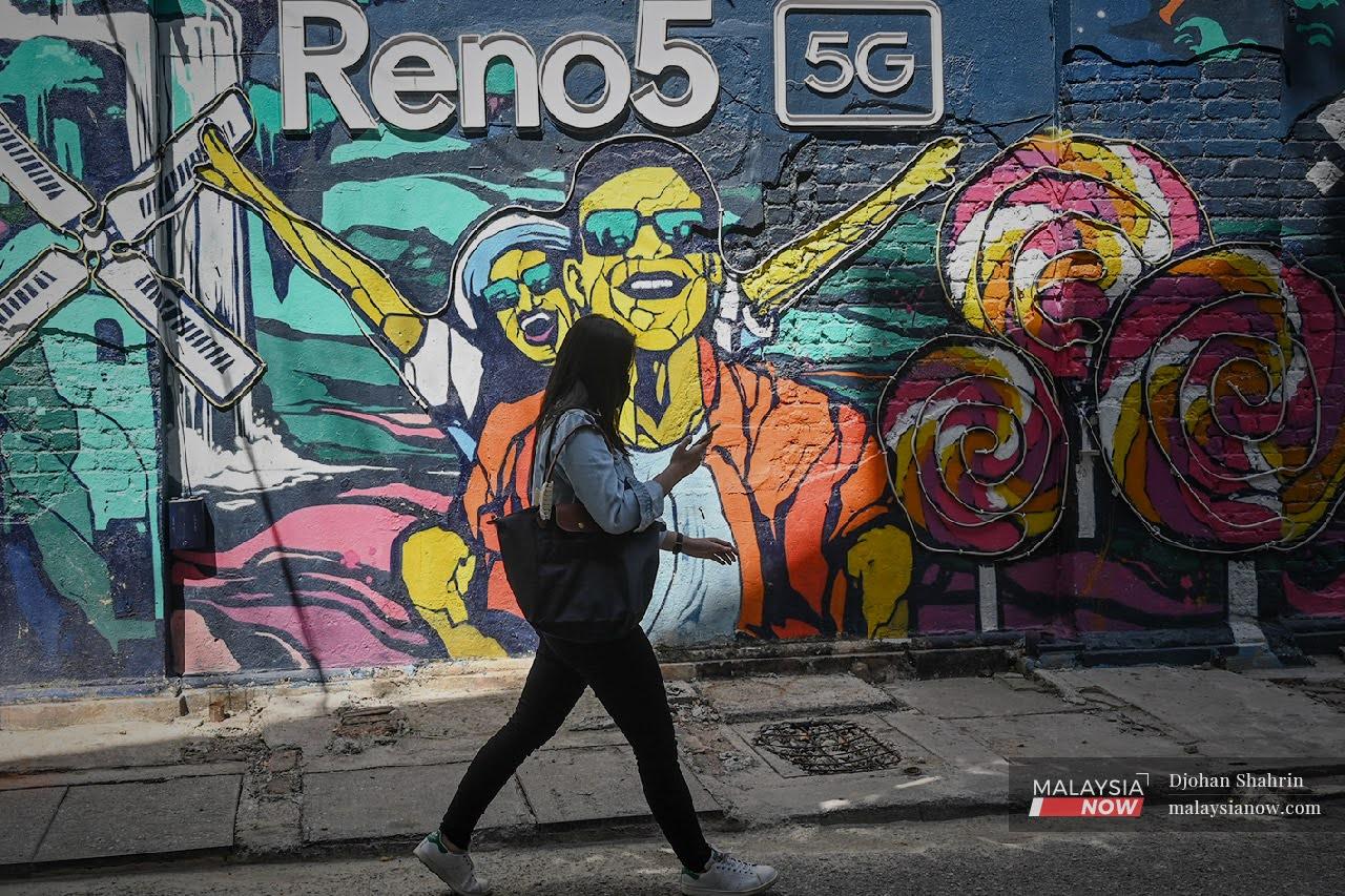 Seorang wanita berjalan melalui sebuah mural sambil melayari internet pada telefon bimbitnya di Jalan Panggung. Orang ramai yang lengkap divaksinasi mendapat kelonggaran untuk mula menjalani kehidupan lebih normal selepas lama menjalani sekatan pergerakan.