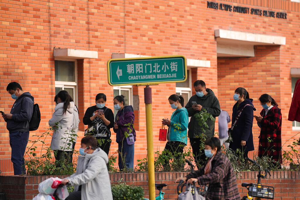 Women wearing face masks to help curb the spread of the coronavirus ride scooters past masked residents linining up to receive booster shots against Covid-19 at a vaccination site in Beijing, Oct 25. Photo: AP