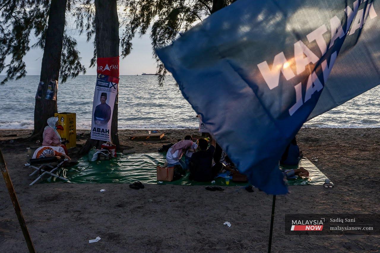 A family picnics on the beach at Pantai Puteri, where Perikatan Nasional flags and posters featuring its chairman Muhyiddin Yassin have been put up.