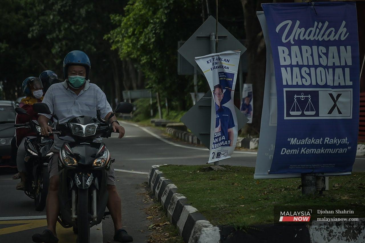 Penunggang motosikal memakai pelitup muka  berhenti di persimpangan jalan yang dipenuhi poster Barisan Nasional di Melaka.