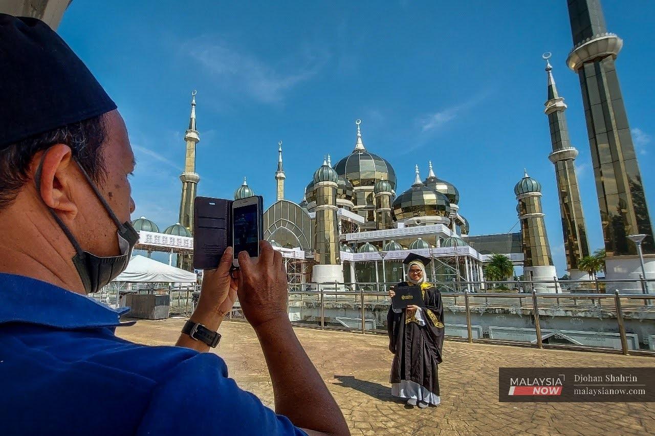Seorang pelawat Masjid Kristal, Terengganu bergambar sambil memakai jubah konvokesyen universiti. Di bawah Pelan Pemulihan Negara, penerima vaksin lengkap diberi kelonggaran untuk melakukan aktiviti ekonomi dan sosial.