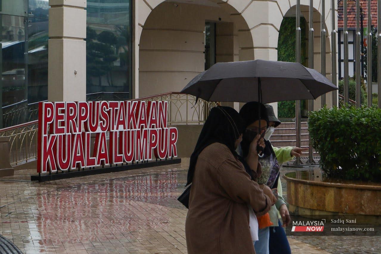 Three women share an umbrella as they make their way through the rain past the Kuala Lumpur Library.