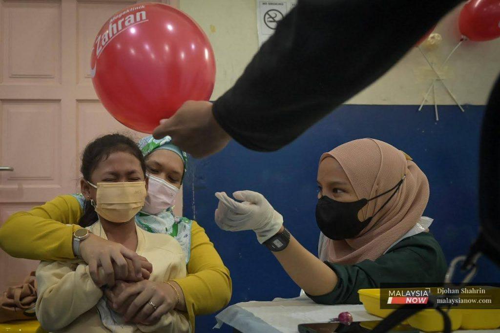 A young girl grimaces as she sits on her mother's lap, waiting for the nurse to administer a dose of Covid-19 vaccine for children as another health worker holds up a balloon to distract her.