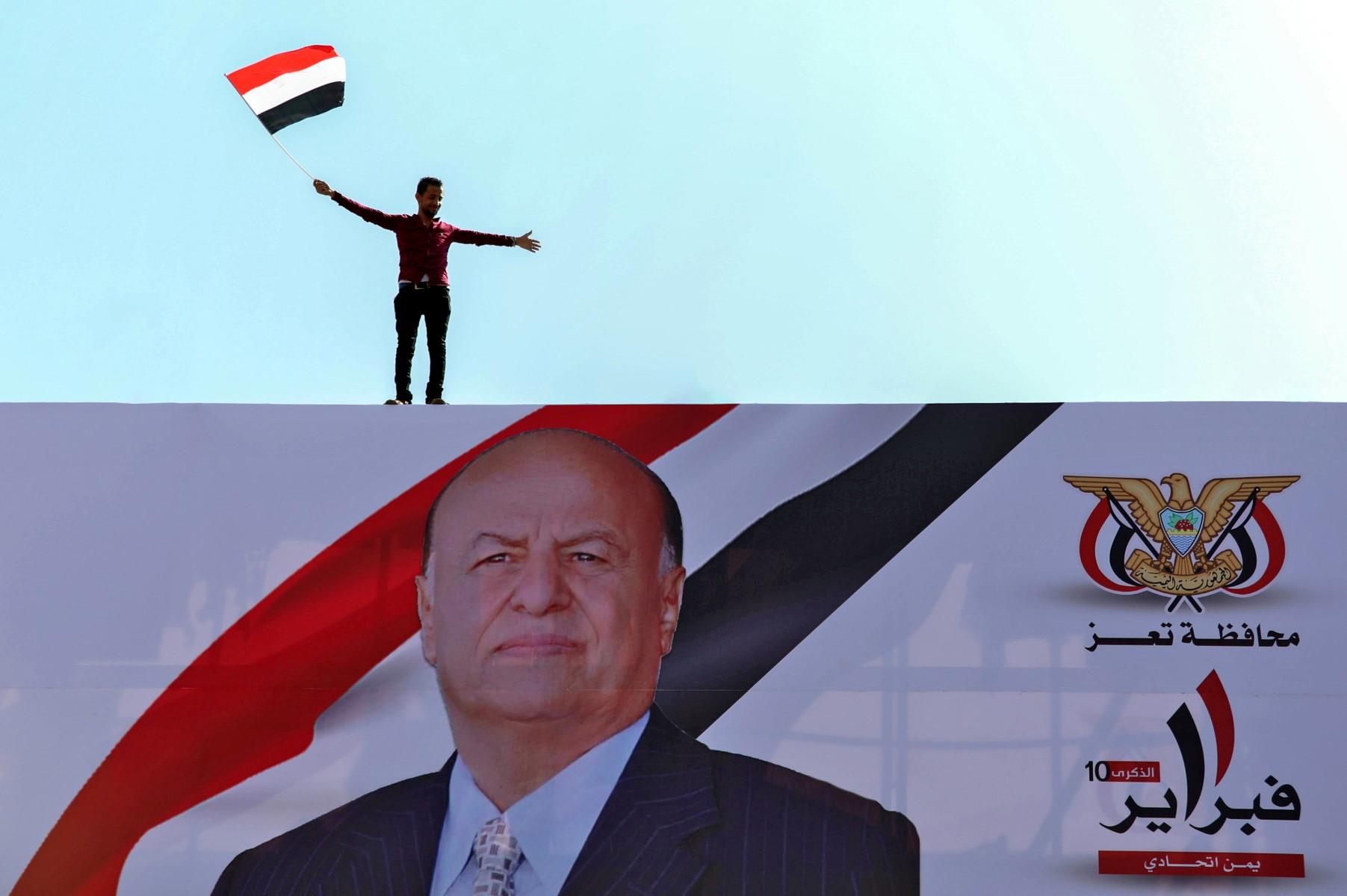 A Yemeni youth waves a national flag atop a billboard showing Yemen's President Abedrabbo Mansour Hadi during a rally commemorating the 10th anniversary of 2011 Arab Spring uprising, in Yemen's third city of Taez on Feb 11, 2021. Photo: AFP