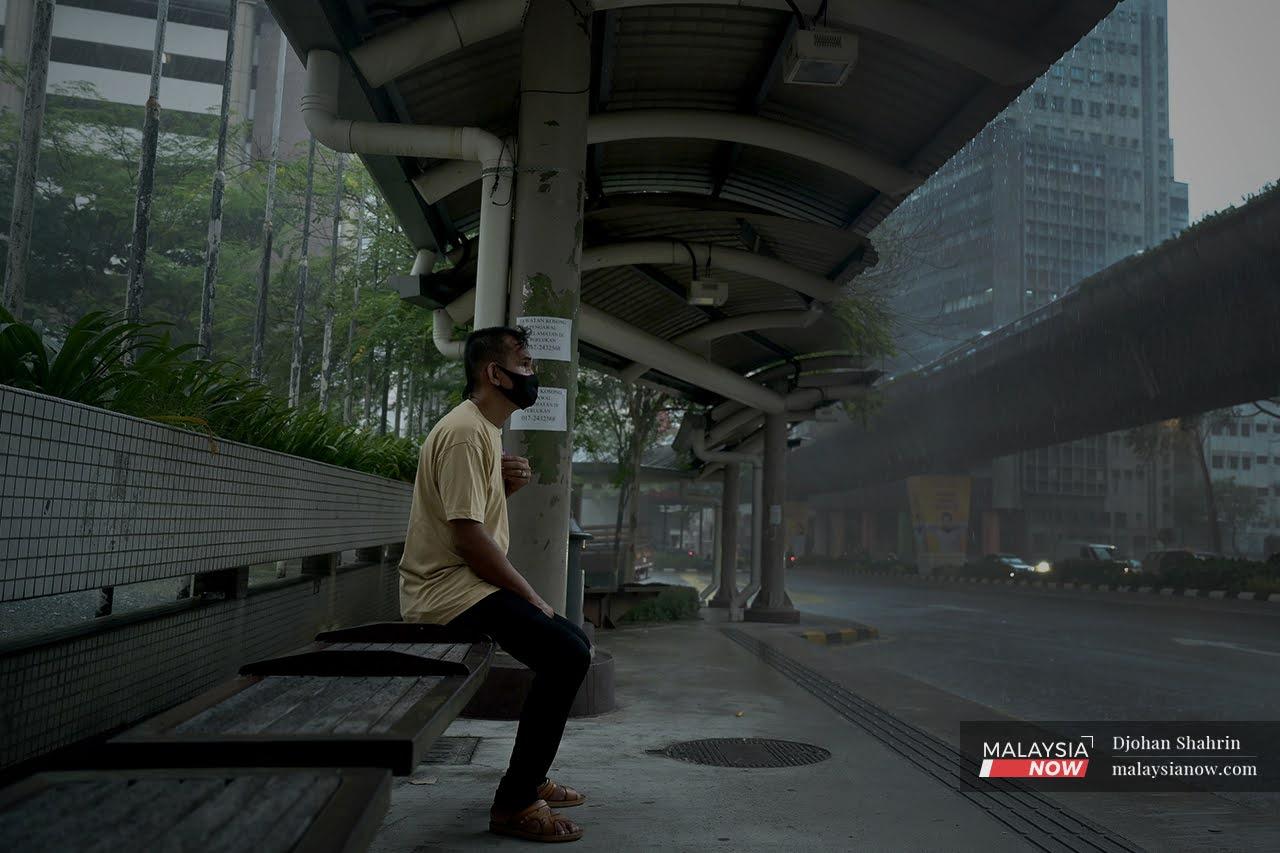 Ruslan sits alone at a bus stop in Kuala Lumpur as rain falls in sheets around him.