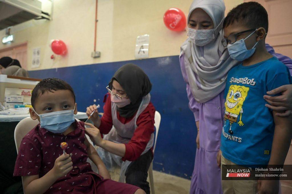 A young boy holds a lollipop in one hand as a nurse administers a dose of Covid-19 vaccine for children while his mother and brother watch, at the Dewan Komuniti Taman Bukit Mewah vaccination centre in Kajang.